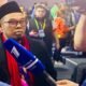Seorang pria berkacamata dengan jas cokelat dan peci hitam berbicara menggunakan mikrofon sambil mengangkat satu jari ke atas, menegaskan pandangannya dalam forum diskusi publik