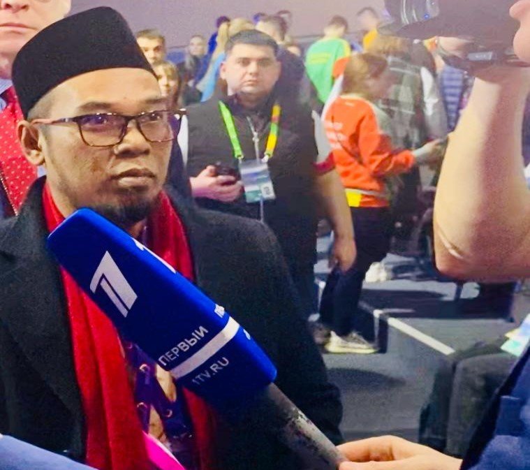 Seorang pria berkacamata dengan jas cokelat dan peci hitam berbicara menggunakan mikrofon sambil mengangkat satu jari ke atas, menegaskan pandangannya dalam forum diskusi publik