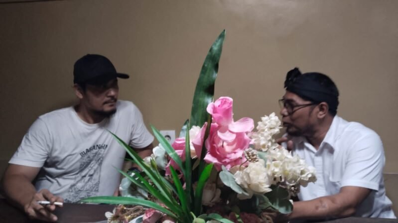 Dua tokoh sedang berdiskusi serius di sebuah meja dengan hiasan bunga di tengahnya; suasana hangat memperlihatkan perbincangan antara aktivis dan tokoh spiritual di Lebak.