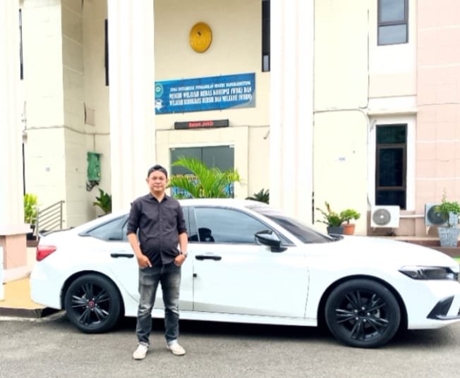 Ketua LBH, Andi Ambrillah,berdiri di depan mobil sedan putih yang terparkir di halaman sebuah gedung pemerintahan.